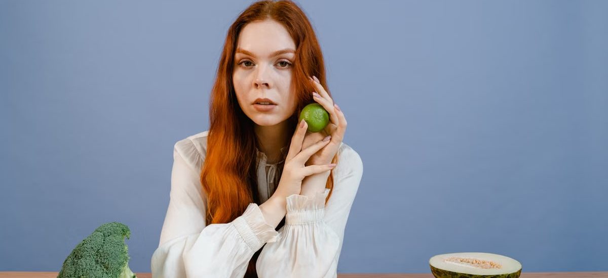 Woman with red hair holding lime surrounded by fresh produce on table.